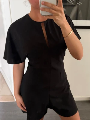 Svart kort jumpsuit med slits - Zara strl XS - Snygg svart kort jumpsuit med vida ärmar och en liten slits vid halsen. Jumpsuiten har en figurnära passform och är tillverkad i ett mjukt, vävt material. Perfekt för dig som gillar stilrena och trendiga plagg. PLAGGET ÄR ALDRIG ANVÄND (lapp kvar)