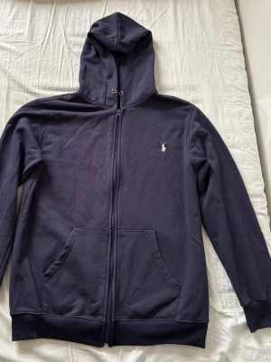 Mörkblå hoodie från Polo Ralph Lauren - Snygg mörkblå hoodie från Polo Ralph Lauren med dragkedja framtill och klassisk logga broderad på bröstet. Hoodien har kängurufickor, ribbade muddar och en skön huva med snörhål. Perfekt för en chill och stilren look.