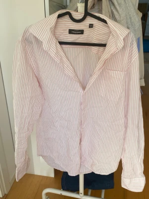 Randig skjorta från Dressmann M - Klassisk skjorta från Dressmann i vitt med tunna röda ränder. Skjortan har lång ärm, bröstficka och knappar framtill. Perfekt för en stilren look och passar till många olika outfits.