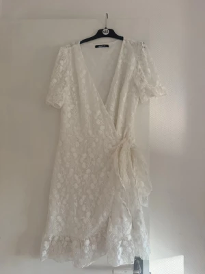 Vit omlottklänning från Ginatricot - Superfin vit omlottklänning med korta ärmar och vackert blommigt spetsmönster. Klänningen har volangkant nertill och knyt i midjan för en feminin siluett. Perfekt för sommarens alla tillfällen!