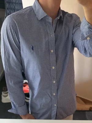 Blårutig skjorta från Ralph Lauren - Klassisk blårutig skjorta från Ralph Lauren med vit knappstängning och broderad logga på bröstet. Skjortan har lång ärm, krage och är tillverkad i bomull. Perfekt för en stilren look. Storlek 38 vilket motsvarar m typ