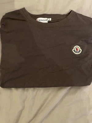 Brun/Grå Moncler t-shirt med logga - Snygg brun/grå t-shirt från Moncler med klassisk rund hals och ikonisk logga på bröstet. Enkel och stilren design som passar perfekt till jeans eller shorts. Tillverkad i mjuk bomull för skön känsla hela dagen. Det står S men skulle säga att den mer passar xs. Pris kan diskuteras vid snabb affär var inte rädd för frågor🤝