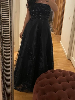 Mörkblå axelbandslös balklänning  - Elegant mörkblå axelbandslös långklänning med glittriga paljettdetaljer och blommiga applikationer. Klänningen har en utsvängd kjol och en vacker, dekorerad överdel som ger en lyxig känsla. Perfekt för speciella tillfällen.