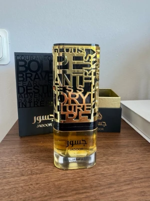 Lattafa Jasoor Eau de Parfum - Lattafa Jasoor Eau de Parfum med en unik, hög cylinderformad flaska i guld och svart med textdetaljer. Doften kommer i en elegant svart och guld presentförpackning med moderna grafiska element. Perfekt för dig som gillar exklusiva och stilrena parfymer.