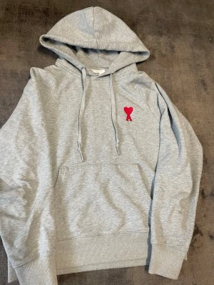 Grå hoodie från Ami Paris med röd logga - Snygg grå hoodie från Ami Paris med klassisk huva och dragsnören. På bröstet sitter Ami:s ikoniska röda hjärta med A-bokstav. Hoodien har magficka och ribbade muddar. Perfekt för en chill och stilren look. Obs! Tredje bilden, visar fläckarna som är på ryggen.