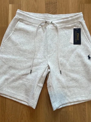 Grå shorts från Polo Ralph Lauren - Snygga ljusgrå shorts från Polo Ralph Lauren med dragsko i midjan och fickor på sidorna samt en bakficka. Mjukt material i bomull, perfekt för sommardagar. Klassisk logga broderad framtill.