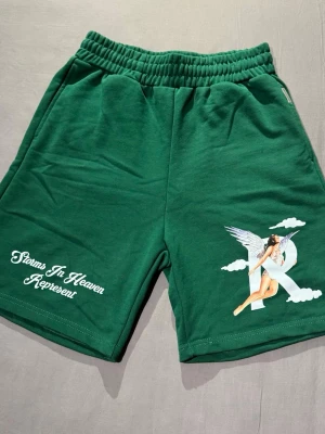 Gröna REPRESENT shorts - Snygga gröna shorts från REPRESENT med elastisk midja och tryck framtill med texten 'Storms In Heaven Represent' samt en stor ängelgrafik. Mjukt material och bakficka för extra stilpoäng. Perfekta för en chill och trendig look. Storlek: M. Använda cirka en gång. 