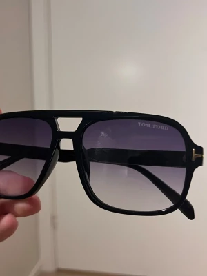 Svarta Tom Ford solglasögon - Snygga svarta solglasögon från Tom Ford med markerad båge och gradienttonade glas. Modellen har en cool, fyrkantig form och detaljer i guld på skalmarna. Perfekt accessoar för att lyfta din stil.