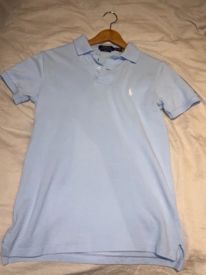Ljusblå pikétröja från Ralph Lauren - Klassisk ljusblå pikétröja från Ralph Lauren i slim fit-modell. Tröjan har korta ärmar, krage med knappar och den ikoniska broderade loggan på bröstet. Tillverkad i mjuk bomull som känns skön mot huden.