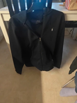 Svart hoodie från Polo Ralph Lauren - Snygg svart hoodie från Polo Ralph Lauren med dragkedja framtill och klassisk vit logga på bröstet. Hoodien har huva och fickor på sidorna. Tillverkad i mjukt bomullsmaterial som är skönt att bära.