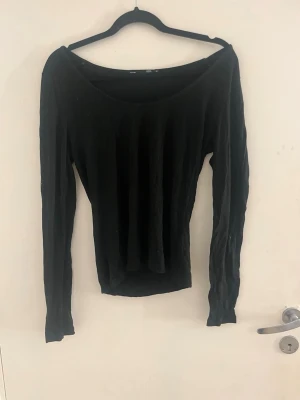 Svart långärmad topp med bred urringning - En svart, figurnära långärmad topp med bred urringning som ger en offshoulder-look. Perfekt att styla med jeans eller kjol för en trendig vibe. Materialet ser ut att vara mjukt och stretchigt, vilket gör den bekväm att bära hela dagen.