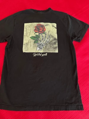 Svart t-shirt Wasted Youth med ros - Svart t-shirt från Wasted Youth med tryck av en skelett-hand som håller en röd ros på ryggen och liten ros på bröstet. Klassisk rund hals och korta ärmar. Perfekt för dig som gillar streetwear och coola prints.