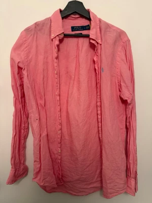 Rosa skjorta från Polo Ralph Lauren - Klassisk rosa skjorta från Polo Ralph Lauren med button-down krage och broderad logga på bröstet. Skjortan har långa ärmar, knappar framtill och är tillverkad i bomull. Perfekt för en avslappnad men ändå stilren look.