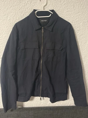Svart overshirt från Lyle & Scott - Stilren svart overshirt från Lyle & Scott med dragkedja framtill och två bröstfickor med lock. Overshirten har klassisk krage och är tillverkad i ett lätt, strukturerat tyg som ger en modern look. Perfekt att bära över en t-shirt för en avslappnad vår stil.