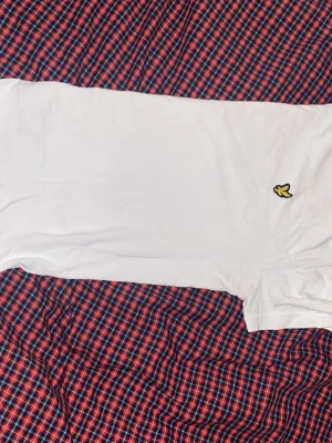 Vit t-shirt från Lyle & Scott - Klassisk vit t-shirt från Lyle & Scott med den ikoniska gula loggan broderad på bröstet. T-shirten har rund hals och korta ärmar, tillverkad i mjuk bomull för en skön känsla. Perfekt basplagg till garderoben. Har inte används så mycket så är i bra skick! 