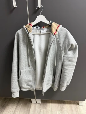 Grå hoodie från Burberry med mönstrad huva - Snygg grå zip-hoodie från Burberry med klassiskt rutmönstrat foder i huvan. Hoodien har dragsko, två fickor framtill och ribbade muddar. Perfekt för en stilren och avslappnad look. Den passar till S/M