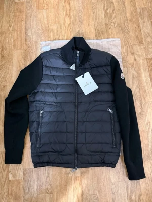 Moncler cardigan  - Fick den idag den är oanvänd helt ny.Snygg svart jacka från Moncler med quiltad front och stickade ärmar. Jackan har hög krage, två dragkedjefickor och Moncler-logga på ärmen. Perfekt för dig som vill ha en stilren och exklusiv look. 