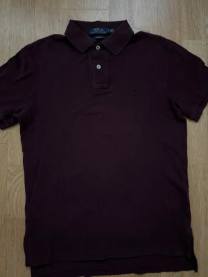 Mörklila pikétröja från Ralph Lauren - Säljer en mörklila pikétröja från Polo Ralph Lauren i modellen Custom Fit. Tröjan har klassisk krage, korta ärmar och knappar framtill. Diskret logga broderad på bröstet. Perfekt för en stilren och avslappnad look.