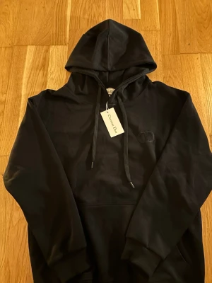 Svart hoodie från Christian Dior - Snygg svart hoodie från Christian Dior med diskret broderad logga på bröstet och klassisk känguruficka. Hoodien har justerbar dragsko i huvan och ribbade muddar. Perfekt för en clean och stilren look.