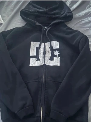 Svart DC zip-hoodie med tryck - Svart hoodie med dragkedja från DC Shoes. Stor vit DC-logga med stjärna på bröstet. Huva med dragsko och två fickor framtill. Perfekt för en avslappnad streetwear-look.