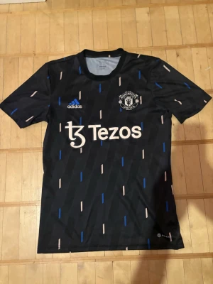 Manchester United svart matchtröja - Snygg svart Manchester United matchtröja från Adidas med vita och blå detaljer samt Tezos-logga på bröstet. Tröjan har rund hals, korta ärmar och är tillverkad i ett lätt och ventilerande material. Perfekt för fotbollsträning eller som supporterplagg.