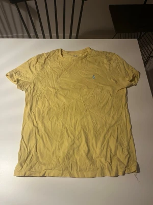 Gul t-shirt från Ralph Lauren - Gul t-shirt från Ralph Lauren med klassisk blå broderad logga på bröstet. T-shirten har rund halsringning och korta ärmar. Tillverkad i mjuk bomull som känns skön mot huden. Perfekt för dig som gillar stilrena och tidlösa plagg.