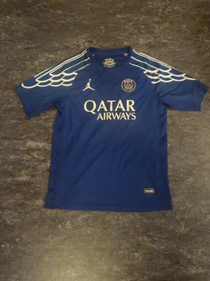 Blå PSG matchtröja Dembele #10 - Snygg blå PSG matchtröja med vita detaljer och vingmönster på axlarna. Framsidan har Qatar Airways-logga och PSG-emblem, baksidan har O. Dembélé och nummer 10 tryckt. Tillverkad i lätt och ventilerande material, perfekt för fotboll eller träning.