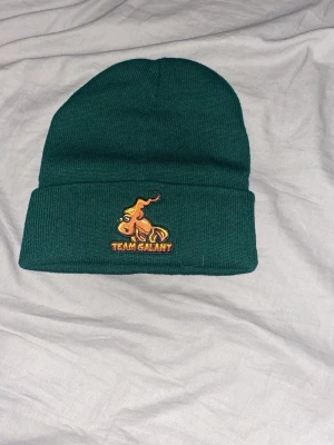 Grön Team Galant mössa - Mörkgrön ribbstickad mössa med uppvikt kant och broderad Team Galant-logga framtill i orange och gult. Perfekt accessoar för dig som gillar streetwear och vill sticka ut med en unik detalj.