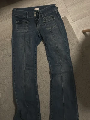 Blå bootcut jeans med fickor fram - Säljer ett par blå bootcut jeans med dubbla framfickor och markerade sömmar. Jeansen har hög midja och stängs med två knappar och dragkedja. Perfekta för en avslappnad och trendig look.