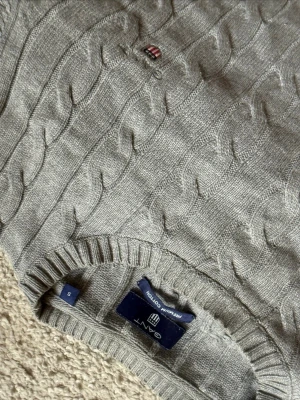 Grå kabelstickad tröja från Gant - Kabelstickad grå tröja från Gant i mjuk bomull. Klassisk rund halsringning och ribbade muddar. Liten broderad Gant-logga på bröstet. Perfekt för en stilren och avslappnad look.