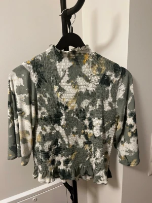 Grön mönstrad smockad topp - Trendig topp med smockad kropp och volangkant nertill. Mönstret är en mix av grönt, vitt och inslag av gult. Toppen har trekvartsärm och lätt rynkad halsringning. Perfekt för dig som gillar unika plagg med coola detaljer.