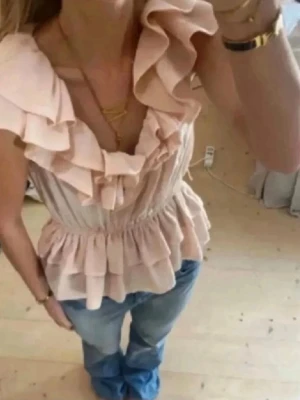 Ljusrosa volangblus med v-ringning - Supersöt ljusrosa topp med volanger och v-ringning. Modellen har breda axelband och smock i midjan som ger en feminin siluett. Perfekt att styla med jeans eller kjol för en trendig look.