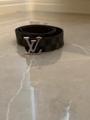  svart rutigt bälte - Snyggt svart bälte med klassiskt rutmönster och en stor silverfärgad LV-logga som spänne. Bältet är gjort i canvas med detaljer i metall och har en modern, stilren look som passar till jeans eller kostymbyxor.