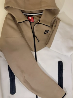 Nike hoodie i beige och vitt - Nike hoodie i snygg beige och vit färgblock-design med svart logga på bröstet. Dragkedja framtill, två svarta dragkedjefickor och huva. Tillverkad i mjukt material, perfekt för en chill och sportig stil.