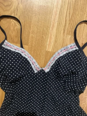 Svart prickig linnetopp med spets - Svart linnetopp med vita prickar och tunna axelband. Toppen har v-ringning med dekorativ spets och rosa längs bysten. Perfekt för en trendig och avslappnad look.