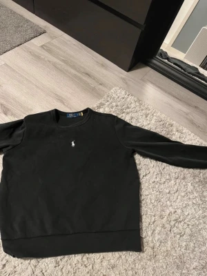Svart sweatshirt från Polo Ralph Lauren - En svart sweatshirt från Polo Ralph Lauren med rund halsringning och den klassiska vita broderade loggan på bröstet. Tröjan har långa ärmar och ribbade muddar vid ärmslut och nederkant. Tillverkad i mjukt bomullsmaterial för en clean och stilren look.