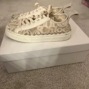 Beige sneakers från Chloé med spetsdetaljer - Snygga beige sneakers från Chloé med spetsdetaljer och tydlig logga på sidan och hälen. Skorna har tjock vit sula, rund tå och breda snören. Materialet är en mix av textil och spets som ger en unik och trendig look. Kommer med original  box och dust bag. Ny pris ca 4500 kr.