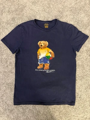 Mörkblå Polo Bear t-shirt Ralph Lauren - Snygg mörkblå t-shirt från Ralph Lauren med ikoniska Polo Bear-trycket på bröstet. Björnen har på sig färgglada kläder och håller en strandboll. Klassisk rund hals och korta ärmar. Perfekt för dig som vill ha en lekfull och stilren look. Längd 53cm midja 45cm