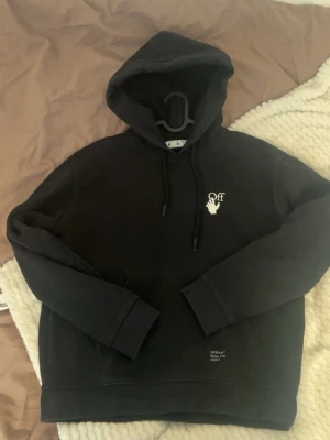 Svart Off-White hoodie med tryck - Säljer en svart hoodie från Off-White med ikoniskt vitt tryck på bröstet och stora pilar på ryggen. Hoodien har huva med dragsko och magficka. Tillverkad i mjukt bomullsmaterial, perfekt för en streetwear-look. Storlek XS men sitter som S-M
