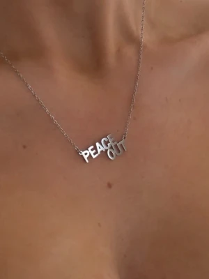 Silverhalsband PEACE OUT ioaku - Snyggt halsband i silverfärgad metall med texten 'PEACE OUT' i stora bokstäver. Kedjan är tunn och enkel, och texten ger ett coolt statement till din stil. Perfekt för dig som vill ha ett personligt och trendigt smycke.