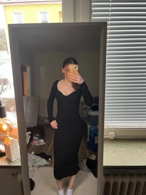 Svart långärmad klänning med v-ringning - Snygg svart långklänning med lång ärm och djup v-ringning. Klänningen har en figurnära passform och är perfekt för dig som gillar en stilren och enkel look. Materialet ser ut att vara stretchigt och bekvämt.