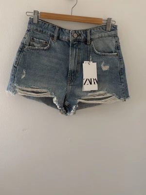 Blå denimshorts från Zara - Snygga blå denimshorts från Zara med hög midja och råa kanter. Shortsen har slitna detaljer framtill och klassiska fem fickor. Perfekta för sommaren och ger en avslappnad vibe till din outfit.