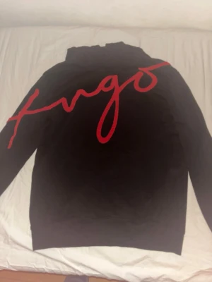 Svart hoodie från Hugo med röd logga - Säljer en svart hoodie från Hugo med stor röd logga i skrivstil över hela framsidan. Hoodien har klassisk huva och ribbade muddar. Materialet känns mjukt och bekvämt, perfekt för en avslappnad streetwear-look.