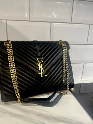 Svart YSL handväska med guldkedja - Säljer en svart handväska från YSL i quiltat läder med ikoniskt guldigt YSL-emblem framtill. Väskan har en elegant guldfärgad kedjerem och klassisk kuvertform. Perfekt för dig som vill ha en lyxig accessoar med statement-detaljer. 