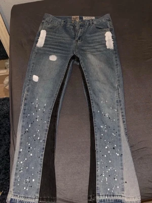 Blå jeans med färgstänk och patchar - Säljer ett par blå jeans med vita färgstänk och slitna patchar på låren. Jeansen har unika svarta paneler längs insidan av benen och en cool, trendig look. Perfekta för dig som vill sticka ut med din stil.
