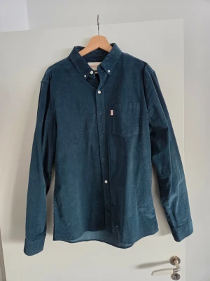 Mörkblå manchesterskjorta Revolution - Snygg mörkblå manchesterskjorta från Revolution med klassisk krage och knappar framtill. Skjortan har en bröstficka och långa ärmar. Perfekt för dig som gillar en stilren och avslappnad look.