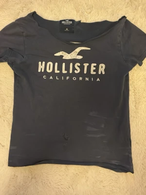Mörkblå Hollister t-shirt XS - Snygg mörkblå t-shirt från Hollister med klassisk logga och texten 'California' framtill. T-shirten har korta ärmar och rund halsringning. Perfekt för en avslappnad och cool stil. Tillverkad i mjuk bomull för skön känsla.