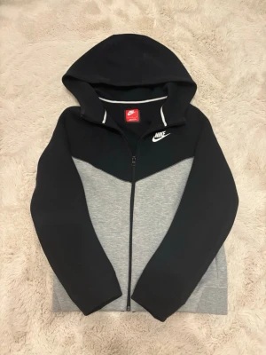 Nike hoodie svart och grå med dragkedja - Nike hoodie i svart och grått med vit logga på bröstet. Tröjan har dragkedja framtill, huva och är tillverkad i mjukt sweatshirtmaterial. Snygg färgblock-design och klassisk sportig look. Perfekt för chill eller träning.