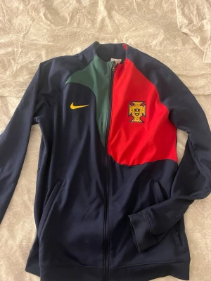 Portugal Nike ziptröja blå/röd - Snygg ziptröja från Nike med Portugals landslagsmärke. Tröjan är mestadels mörkblå med röda och gröna paneler samt gul Nike-logga. Perfekt för dig som gillar fotboll och vill ha en sportig look. Tillverkad i mjukt, stretchigt material.