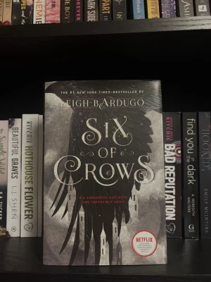 Six of Crows av Leigh Bardugo (Hardcover) - Säljer boken 'Six of Crows' av Leigh Bardugo. Omslaget är svart och grått med en stor svart kråka och siluetter av byggnader. Boken är på engelska och har en Netflix-sticker på framsidan. Perfekt för dig som gillar fantasy och spänning.
